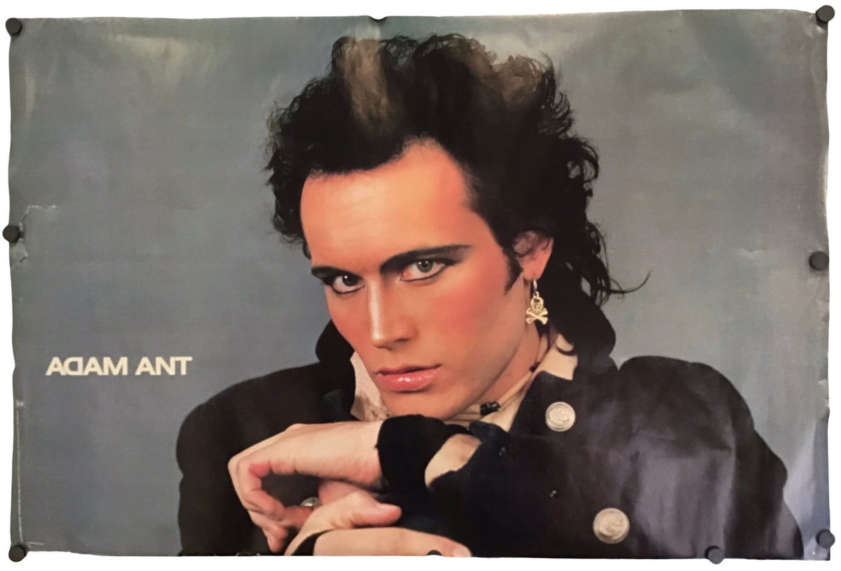 A rectangular vintage poster or print featuring a portrait of Adam Ant against a gray background, with the text 'ADAM ANT' printed on the left side. The print is photographed hanging or mounted with visible clips or fasteners along the left and top edges.