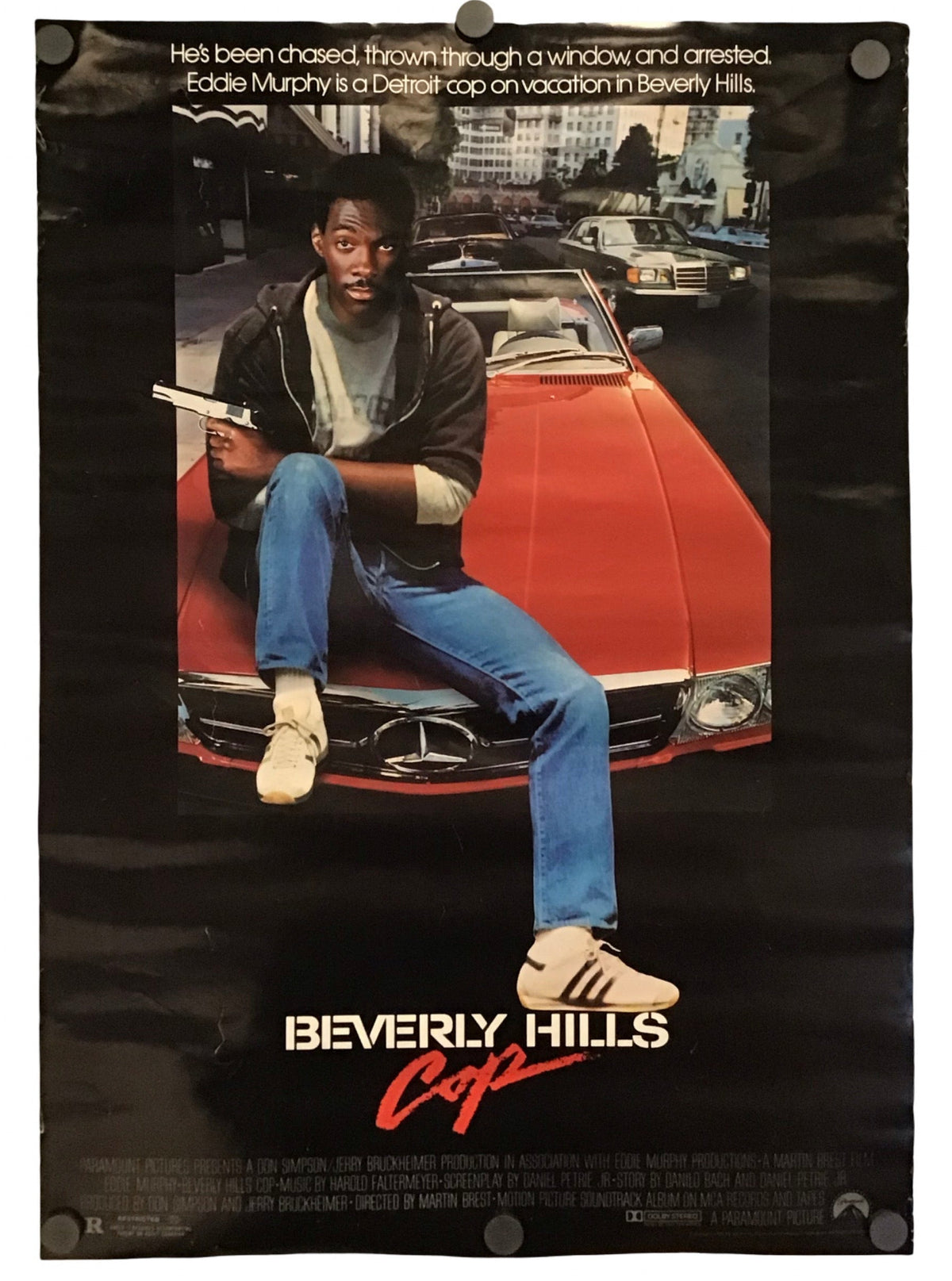 A vintage movie poster for 'Beverly Hills Cop' featuring Eddie Murphy seated on a red car, with the title at the bottom and a black background around the printed artwork.