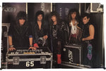 A rectangular printed banner or poster featuring a group of five rock band members standing behind equipment cases. The banner is photographed against a plain white background with a few small detached pieces visible near the edges.