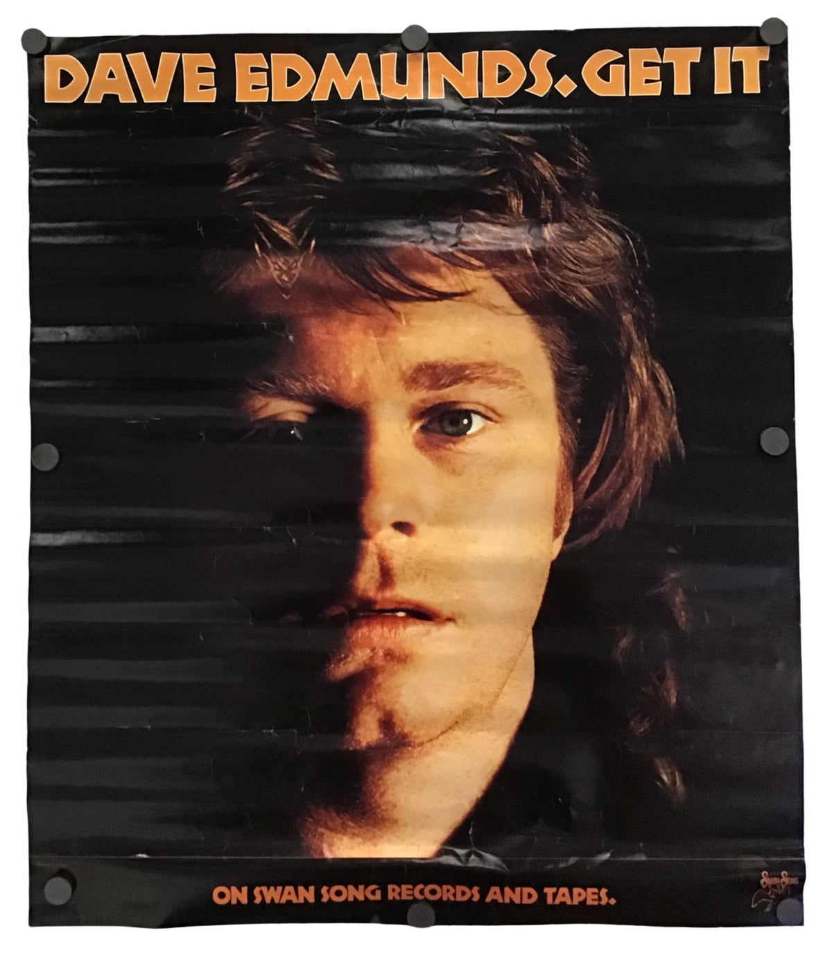 A vintage-style Dave Edmunds 'Get It' poster or album cover hanging against a white wall. The design features a dark portrait of Dave Edmunds with bold orange text reading 'DAVE EDMUNDS GET IT' at the top and additional orange text at the bottom reading 'ON SWAN SONG RECORDS AND TAPEL.
