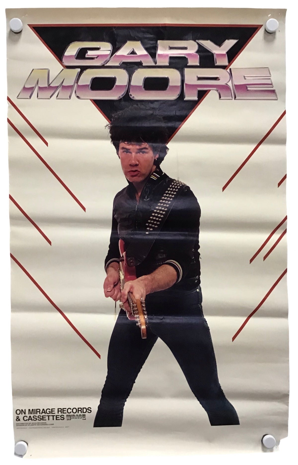 A vintage-style poster for Gary Moore hangs against a white wall, secured with four white binder clips at the corners. The poster features a cream background with red graphic lines and a large black triangular shape behind the text 'GARY MOORE' in metallic pink and silver lettering.