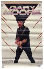 A vintage-style poster for Gary Moore hangs against a white wall, secured with four white binder clips at the corners. The poster features a cream background with red graphic lines and a large black triangular shape behind the text 'GARY MOORE' in metallic pink and silver lettering.
