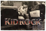 A rectangular music poster for the band Kid Rock, featuring a black-and-white photo of a man in a hat sitting in the back of a truck and holding a guitar, with the text 'KID ROCK' in large patriotic red, white, and blue lettering across the bottom.