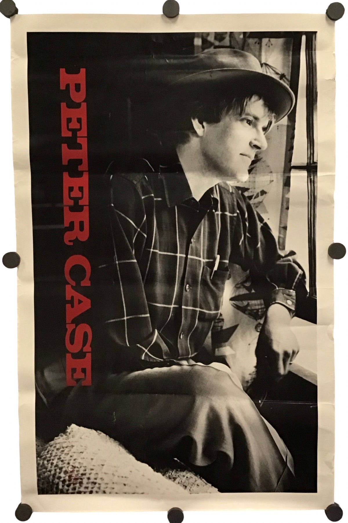 A vintage Peter Case poster featuring a black and white photograph of a man in a hat, with bold red vertical text reading 'PETER CASE' on the left, photographed against a white background with visible mounting clips at the corners.