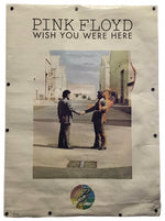 A vintage Pink Floyd 'Wish You Were Here' poster displayed flat against a white background, featuring the band's name and album title at the top, with the iconic handshake photo in the center and a small circular sticker near the bottom.