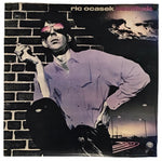 A square album cover for 'Anomalous' by Ric Ocasek, featuring a stylized portrait of a man in a lavender shirt and pink sunglasses against a brick wall and dark night sky.