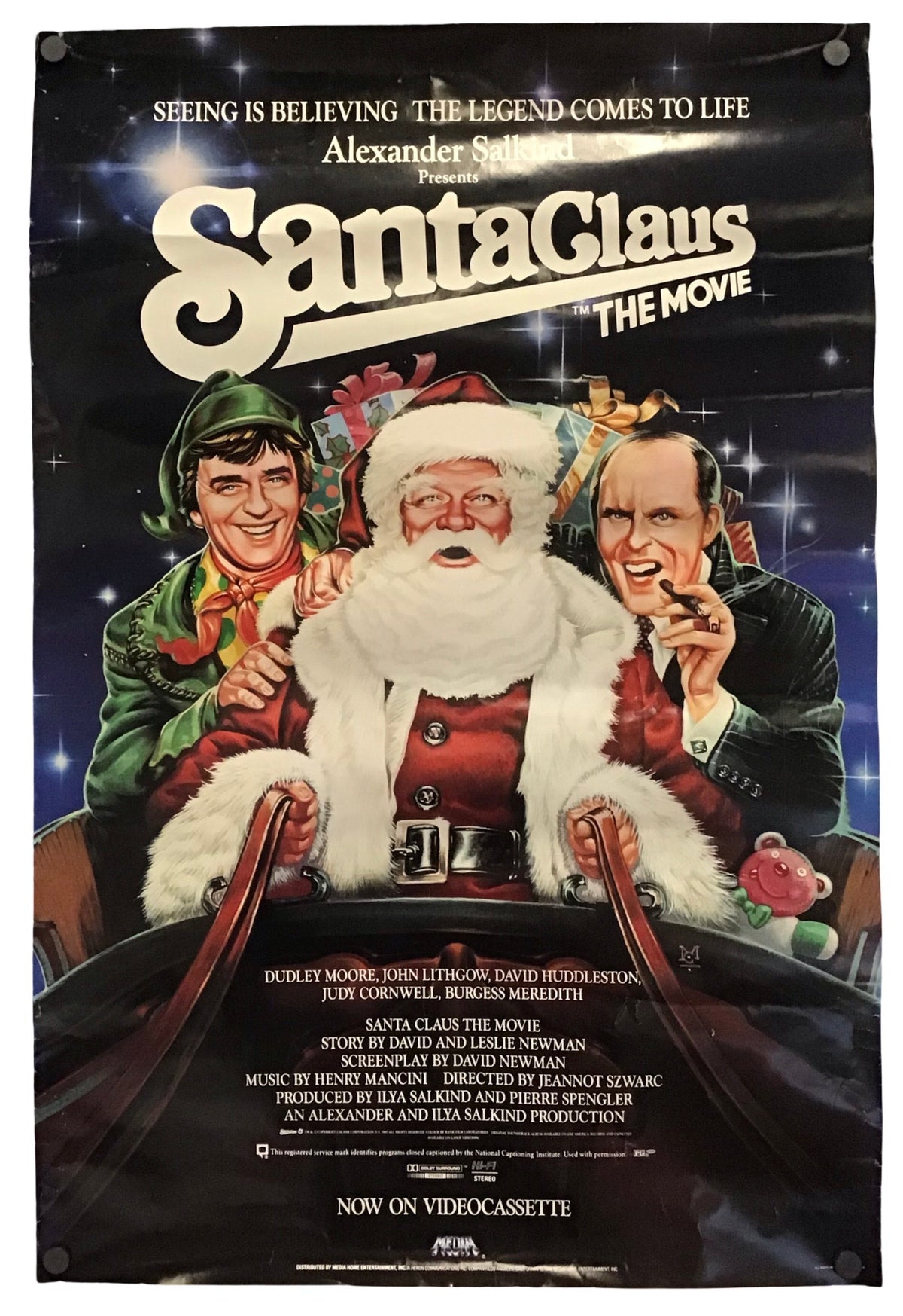 A vintage movie poster for 'Santa Claus The Movie' featuring a colorful illustration of Santa Claus in a sleigh with two men, set against a dark starry background. The poster includes text for the film and credits at the bottom.