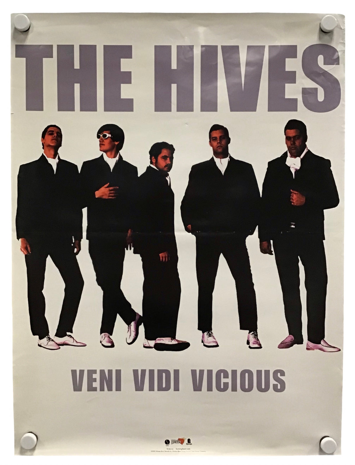 A vintage-style band poster for 'The Hives' featuring five men in black suits against a light background, with the text 'THE HIVES' at the top and 'VENI VIDI VICIOUS' at the bottom. The poster is shown hanging with visible wall fasteners.