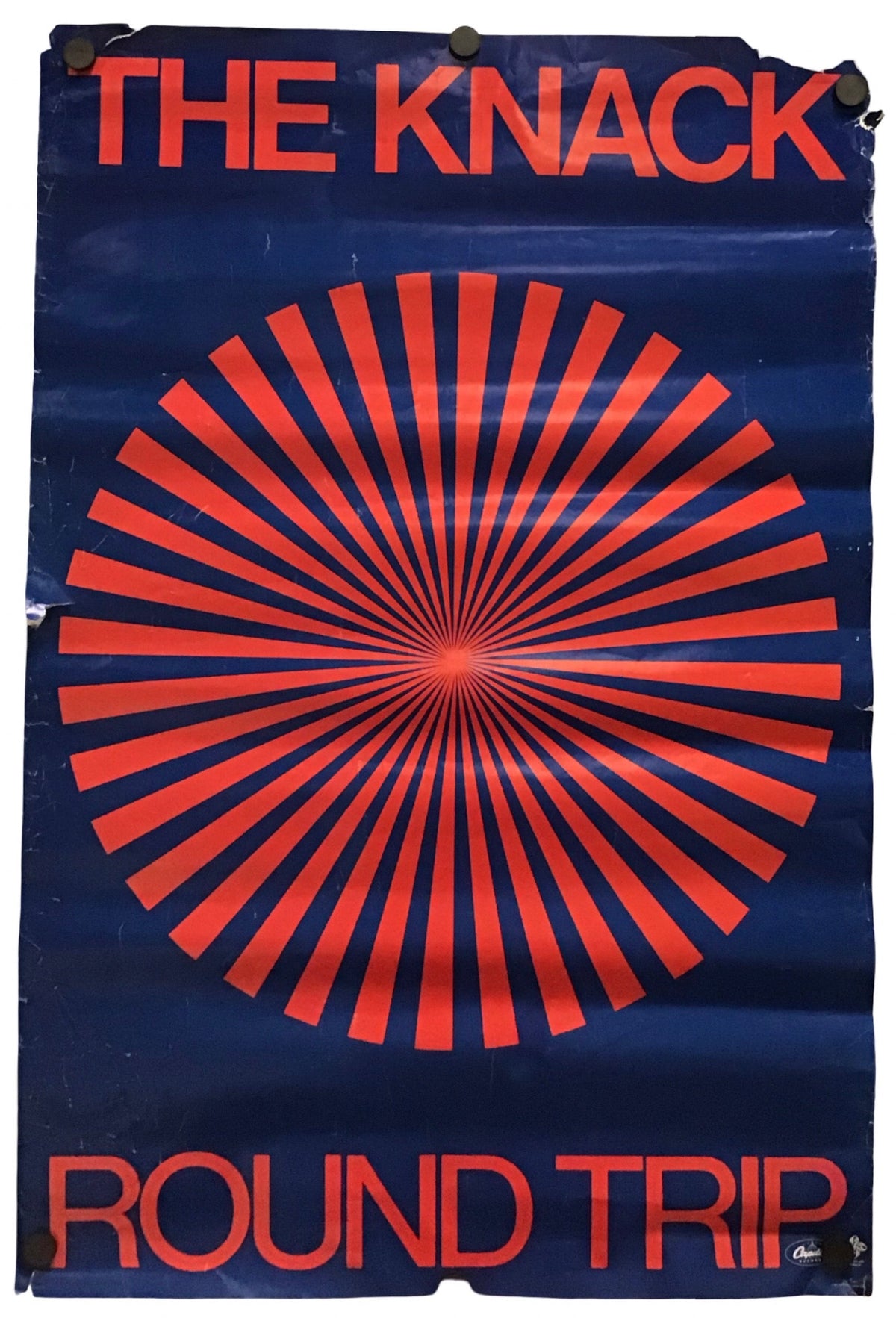 A vintage-style poster or print with the words 'THE KNACK' at the top and 'ROUND TRIP' at the bottom, featuring a large red radial burst on a dark blue background. The poster appears slightly worn at the edges and is photographed against a plain white background.