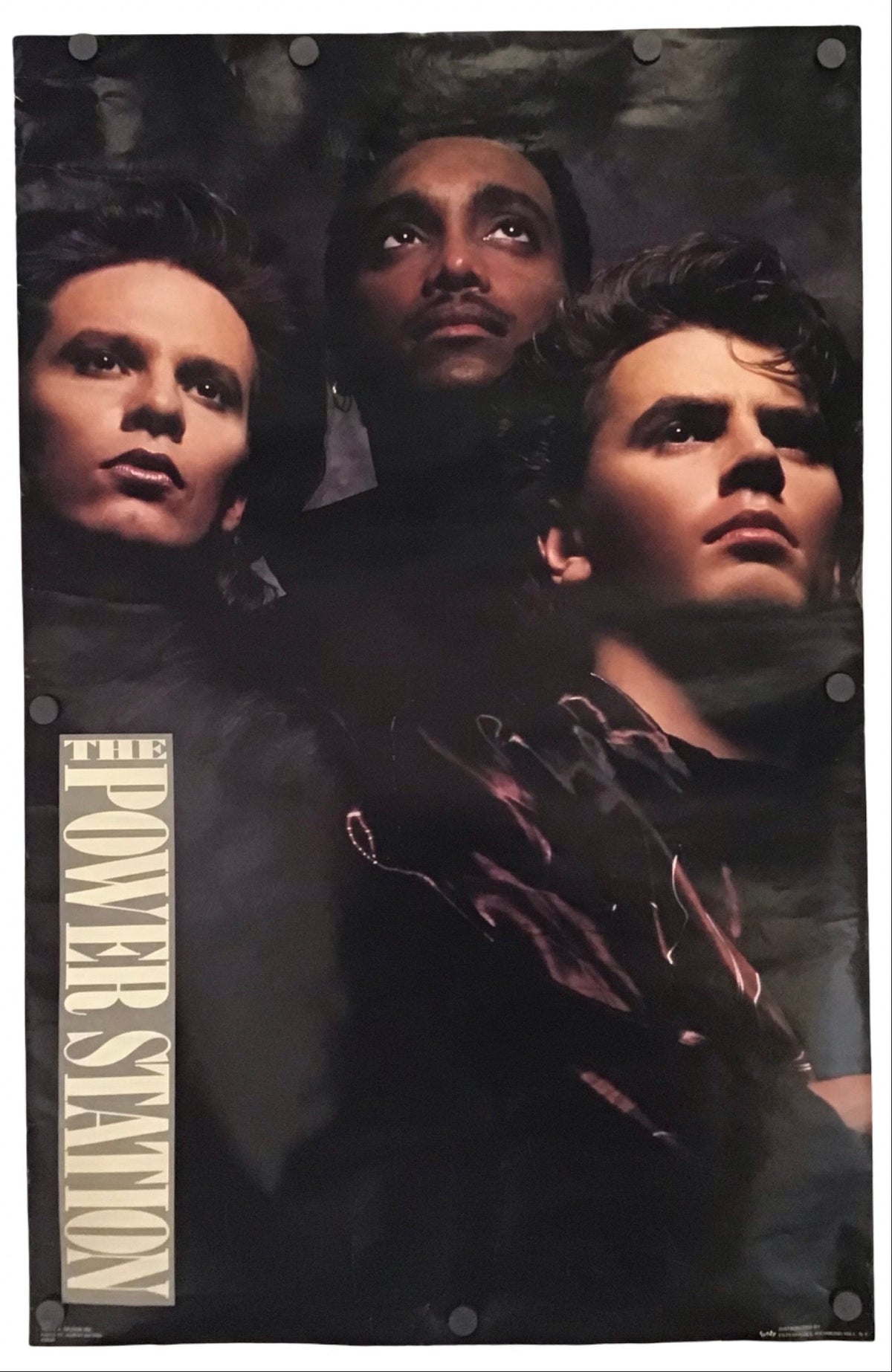 A vertical band poster for 'The Power Station' featuring the three members of the band with their faces partially obscured by shadows and a dark, moody background. The text 'THE POWER STATION' is printed vertically on the left side in a light beige font.