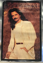 A vintage Yanni 'Dare to Dream' poster featuring the artist with long dark hair and a mustache, wearing a cream-colored outfit against a warm brown background, mounted on a metallic silver wall.