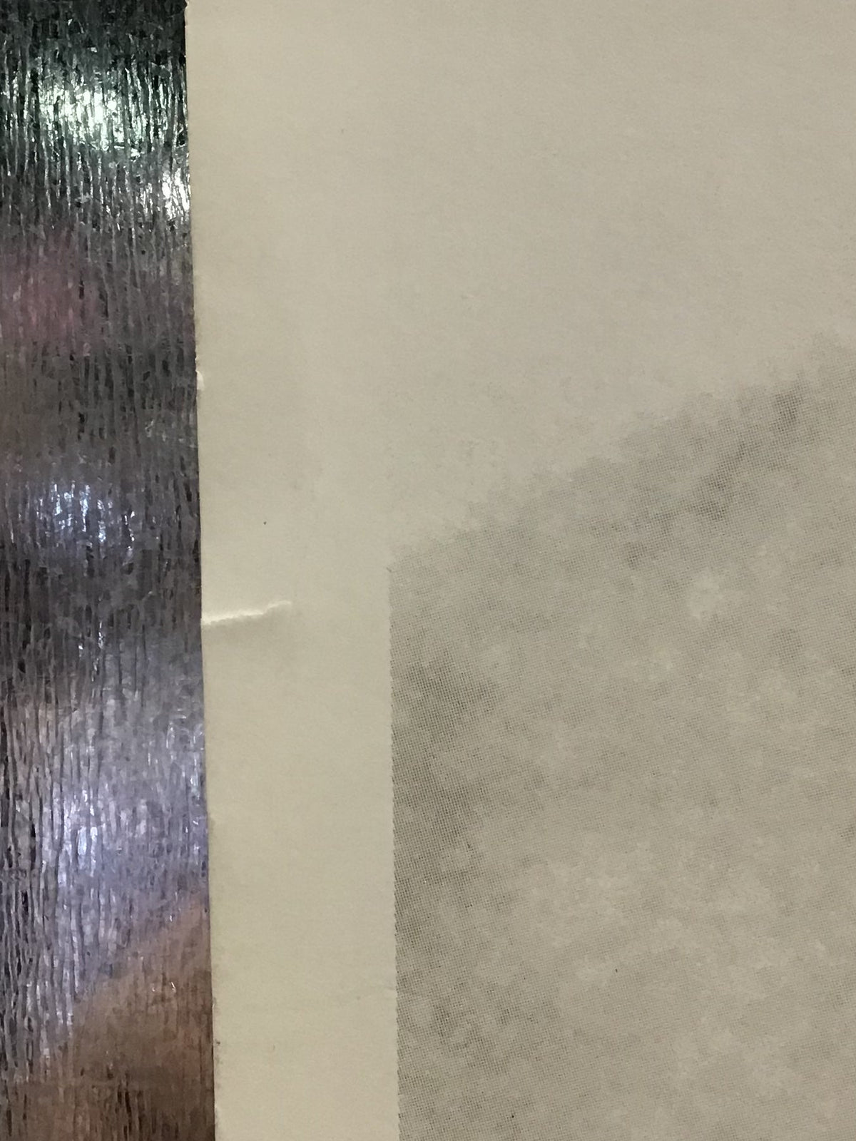 A close-up view of a textured surface with a horizontal seam or fold, showing a light gray upper section and a reflective dark metallic lower section with blurred colored reflections.