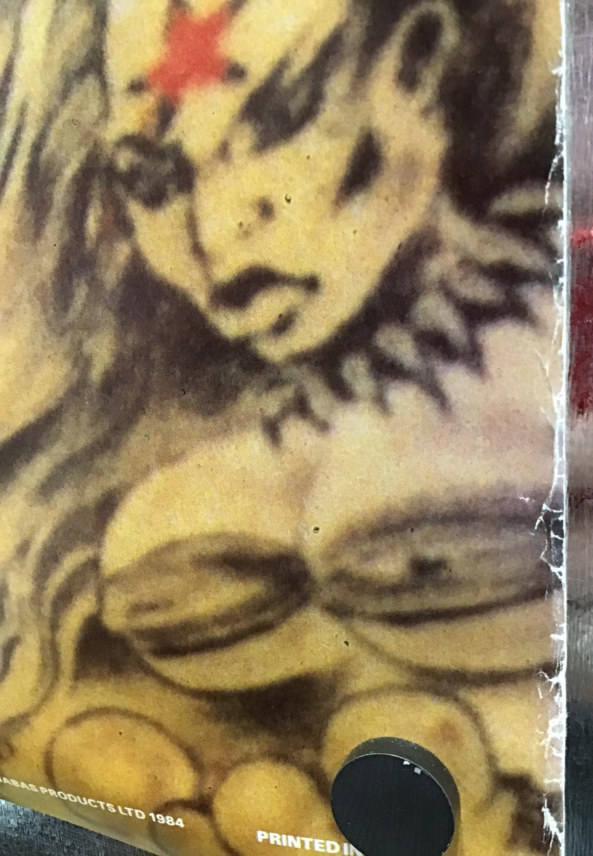 A close-up of a printed graphic featuring a stylized face in black, yellow, and brown tones with a small red star accent, photographed against a dark reflective surface.