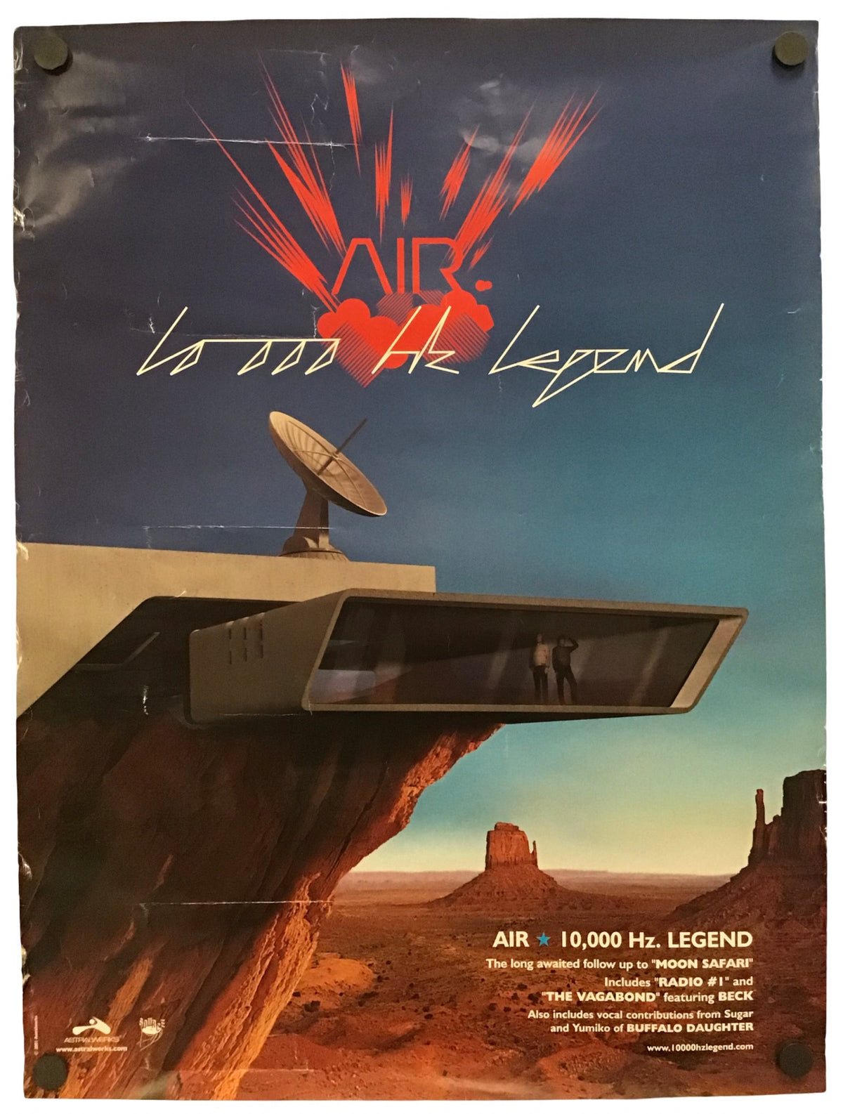 A vintage-style poster or print featuring a desert landscape with a large rock formation, a satellite dish, and bold red/orange graphic text reading 'AIR' and '10 000 Hz Legend' against a blue sky. The poster is photographed hanging against a white wall with clips visible at the top.