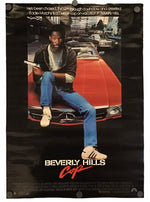 A vintage movie poster for 'Beverly Hills Cop' featuring Eddie Murphy seated on a red car, with the title at the bottom and a black background around the printed artwork.