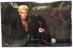 A signed photo print of Billy Idol, featuring the artist in a black leather jacket and brown boots against a dark studio background, with a prominent pink autograph in the upper right corner.
