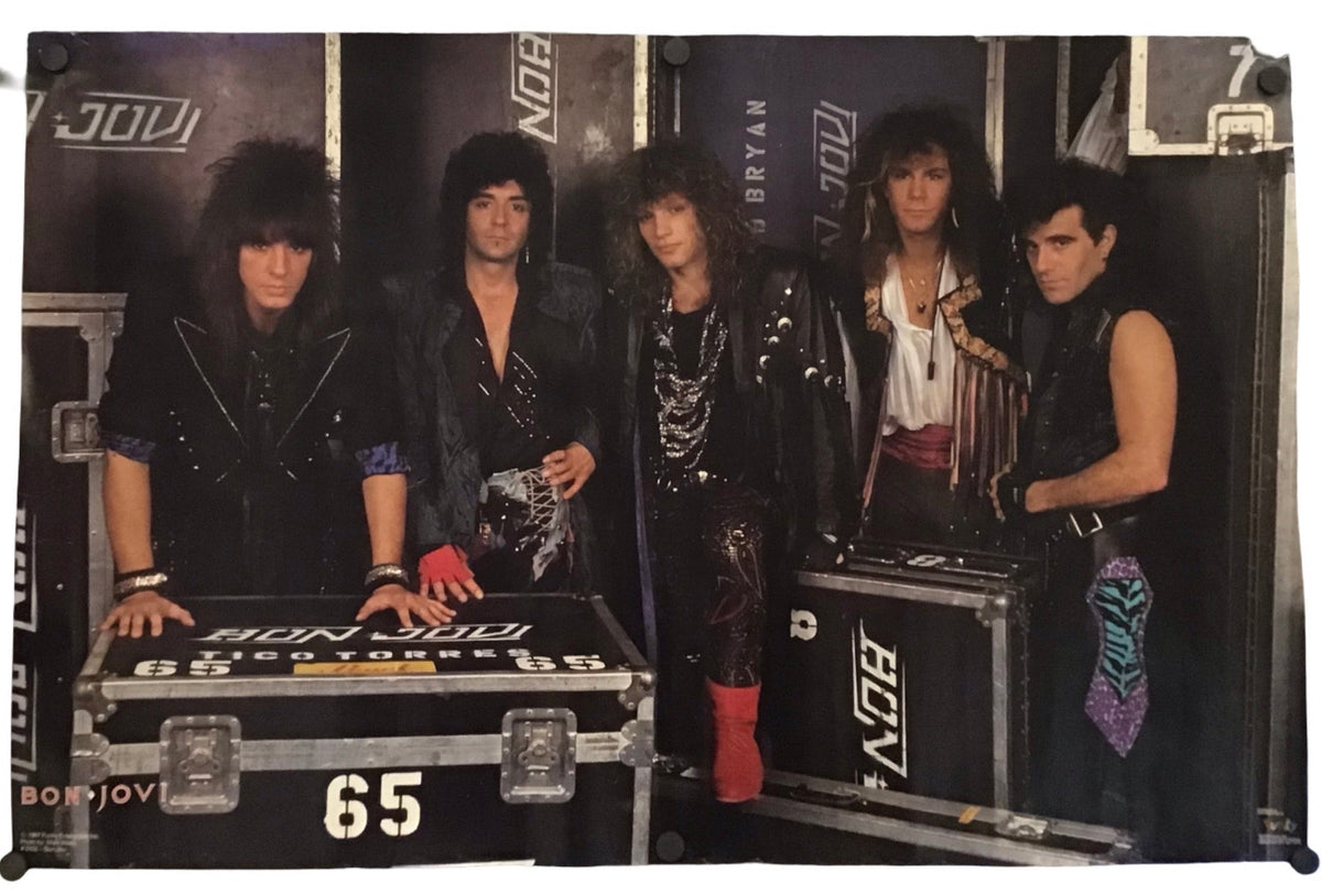 A rectangular printed banner or poster featuring a group of five rock band members standing behind equipment cases. The banner is photographed against a plain white background with a few small detached pieces visible near the edges.