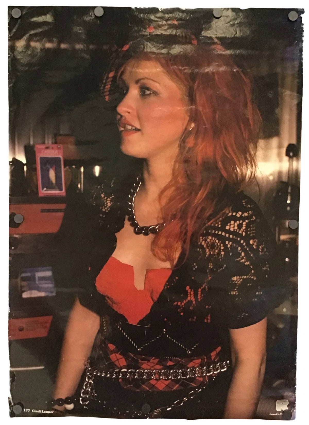 A vertical photographic print or poster showing a woman with red hair wearing a black patterned top over a red dress, photographed against a dark interior background. The print is displayed flat with visible mounting holes along the edges.