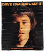 A vintage-style Dave Edmunds 'Get It' poster or album cover hanging against a white wall. The design features a dark portrait of Dave Edmunds with bold orange text reading 'DAVE EDMUNDS GET IT' at the top and additional orange text at the bottom reading 'ON SWAN SONG RECORDS AND TAPEL.