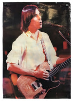 A vertical photographic print or poster showing a male musician with shoulder-length reddish-brown hair singing into a microphone while holding an electric guitar. The print is photographed hanging against a white background with visible pin points at the top corners.