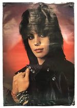 A vintage-style poster or print featuring a person with dark hair, a black leather jacket, and a red-orange gradient background, photographed against a light neutral surface with pins at the corners.