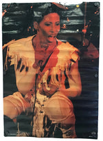A vertical photographic poster or print showing a performer seated and singing into a microphone, with a dark textured background and warm orange-red lighting. The print is photographed hanging against a light gray wall, with visible mounting points and slight glare near the top.