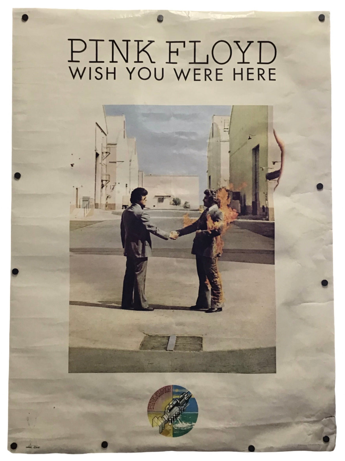A vintage Pink Floyd 'Wish You Were Here' poster displayed flat against a white background, featuring the band's name and album title at the top, with the iconic handshake photo in the center and a small circular sticker near the bottom.