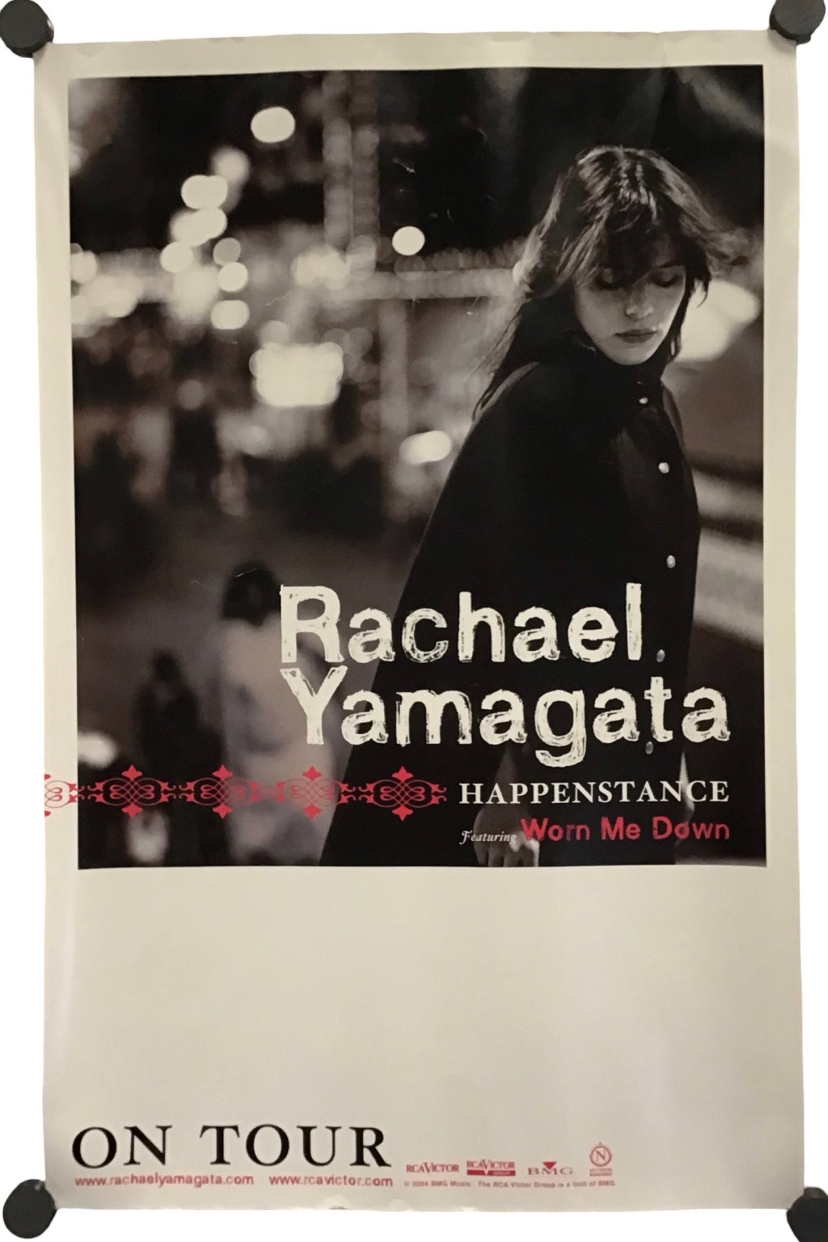 A vertical concert poster for Rachael Yamagata's 'Happenstance' tour, featuring a black and white photograph of the artist, with red and white text, displayed on a white background with black mounting clips at the corners.