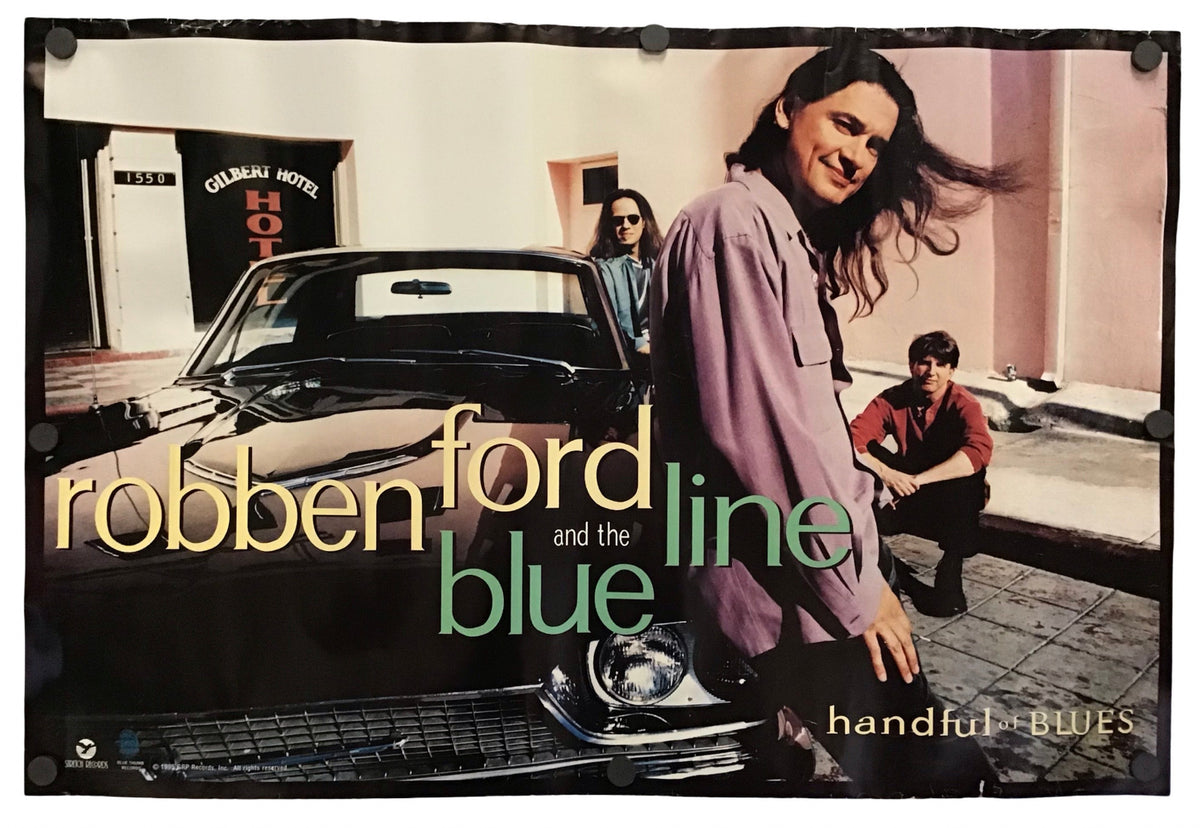 A vintage-style band poster for 'Robben Ford and the Blue Line' hangs against a white wall. The poster features a photograph of three men standing beside a black car, with the band's name and song title 'Handful of Blues' printed prominently in yellow and green text.