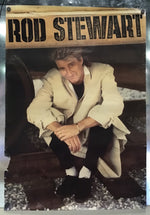 A vintage Rod Stewart poster or print mounted on a metallic surface. The poster features a photo of Rod Stewart crouching in a light beige trench coat, black trousers, and black shoes, with a tan banner at the top reading 'ROD STEWART' in bold black letters.