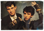 A vintage-style photographic print or poster showing two men in dark suits and accessories against a smoky gray-blue background, photographed against a white backdrop.
