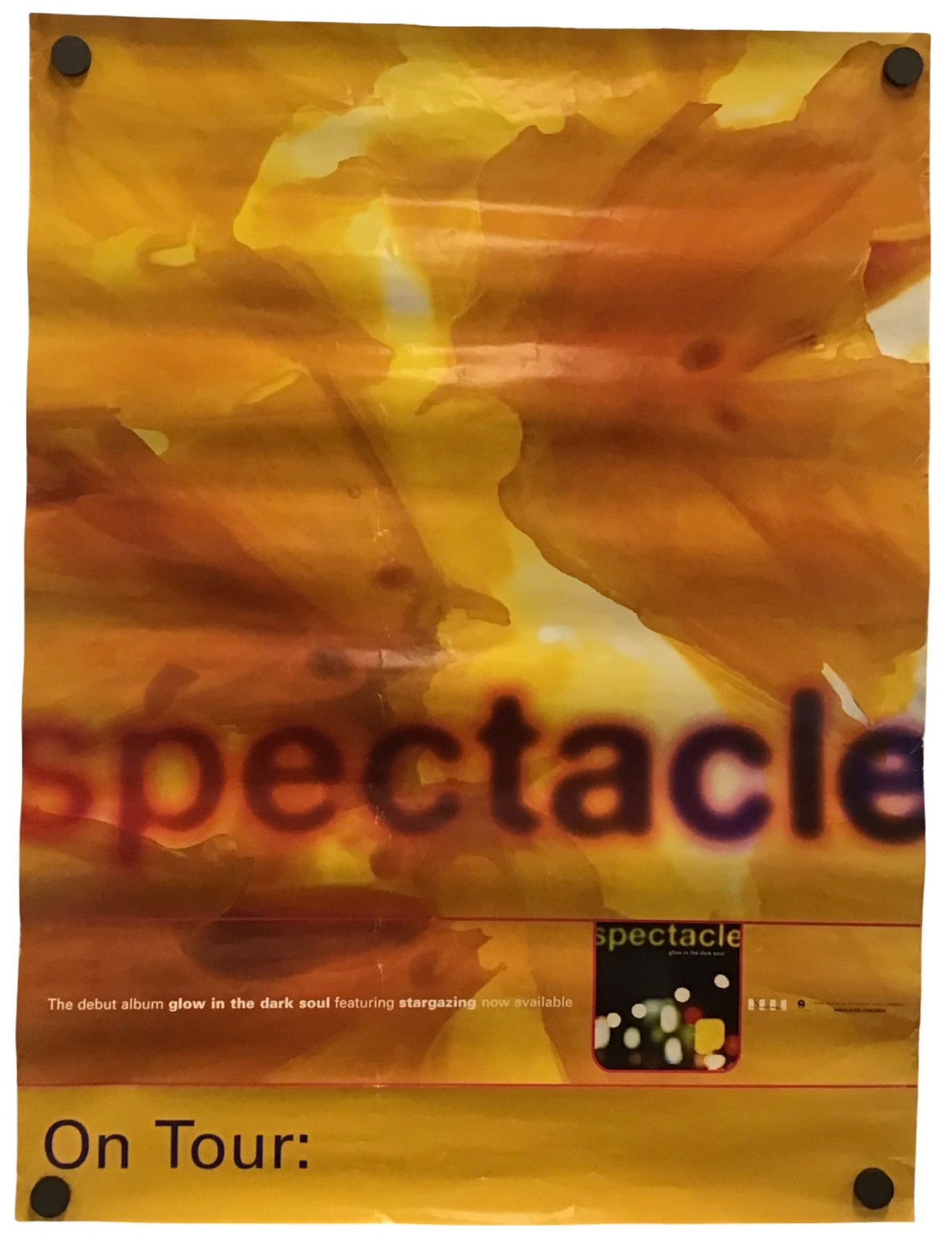 A rectangular yellow and orange abstract poster with the word 'spectacle' across the center and 'On Tour:' at the bottom, photographed against a white background with black binder clips visible at the corners.