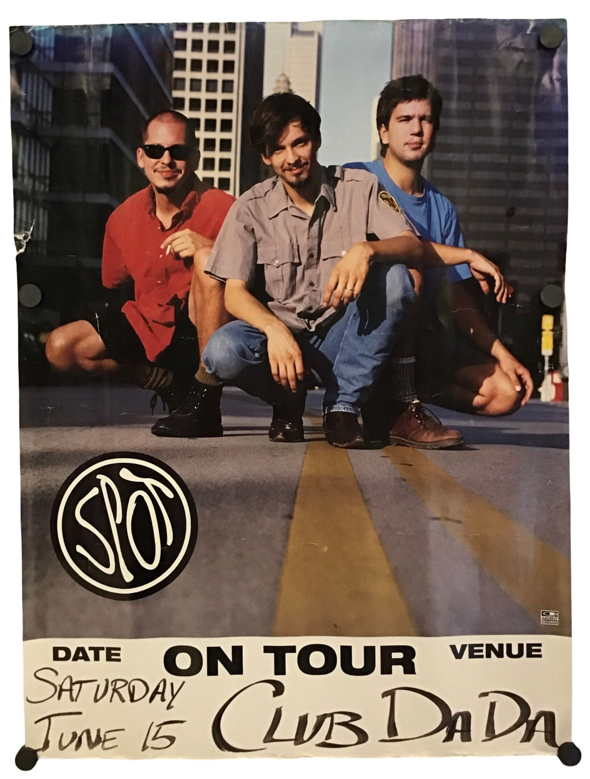 A vintage concert poster for the band Spot, featuring three band members posing on a city street with skyscrapers in the background. The bottom of the poster includes printed text reading 'DATE ON TOUR VENUE Saturday June 15 Club Da Da' and a circular SIO logo.