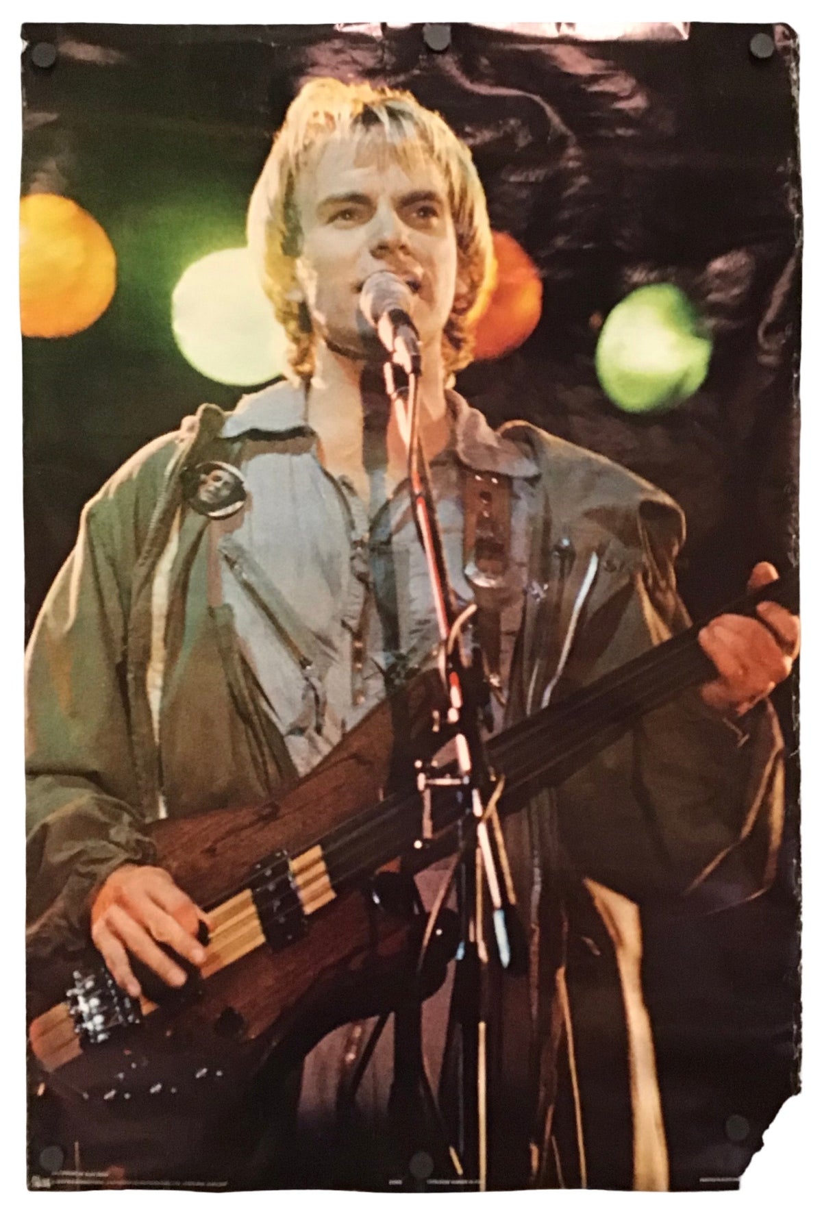 A vertical photographic print or poster showing a male musician performing on stage, holding a bass guitar and singing into a microphone. The print is photographed against a plain white background with visible mounting clips at the top.