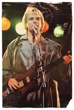 A vertical photographic print or poster showing a male musician performing on stage, holding a bass guitar and singing into a microphone. The print is photographed against a plain white background with visible mounting clips at the top.