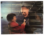 A rectangular printed artwork or poster showing a smiling bald man in a black leather jacket holding a small red-and-white object, with a child in a red jacket reaching toward him. The print is photographed against a plain white background and appears to be mounted or photographed flat with visible round fasteners along the left edge.