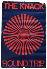 A vintage-style poster or print with the words 'THE KNACK' at the top and 'ROUND TRIP' at the bottom, featuring a large red radial burst on a dark blue background. The poster appears slightly worn at the edges and is photographed against a plain white background.
