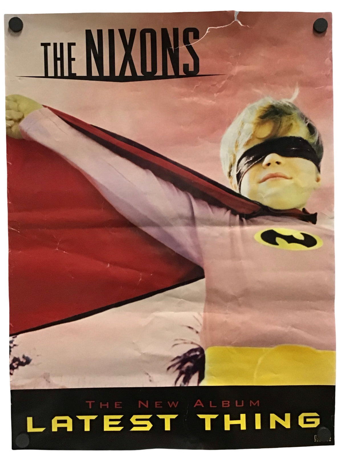 A vintage-style poster or album cover featuring the text 'THE NIXONS' at the top and 'THE NEW ALBUM LATEST THING' at the bottom, with a child in a superhero costume pulling a red cape across a pink background. The poster is photographed hanging against a white wall with two black wall hooks visible.