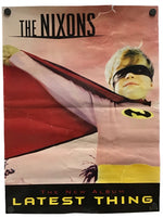 A vintage-style poster or album cover featuring the text 'THE NIXONS' at the top and 'THE NEW ALBUM LATEST THING' at the bottom, with a child in a superhero costume pulling a red cape across a pink background. The poster is photographed hanging against a white wall with two black wall hooks visible.