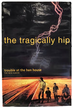 A rectangular album poster for 'the tragically hip' featuring a dark stormy sky with lightning, yellow title text, and a desert road scene with figures at the bottom. The poster is photographed against a plain white background.