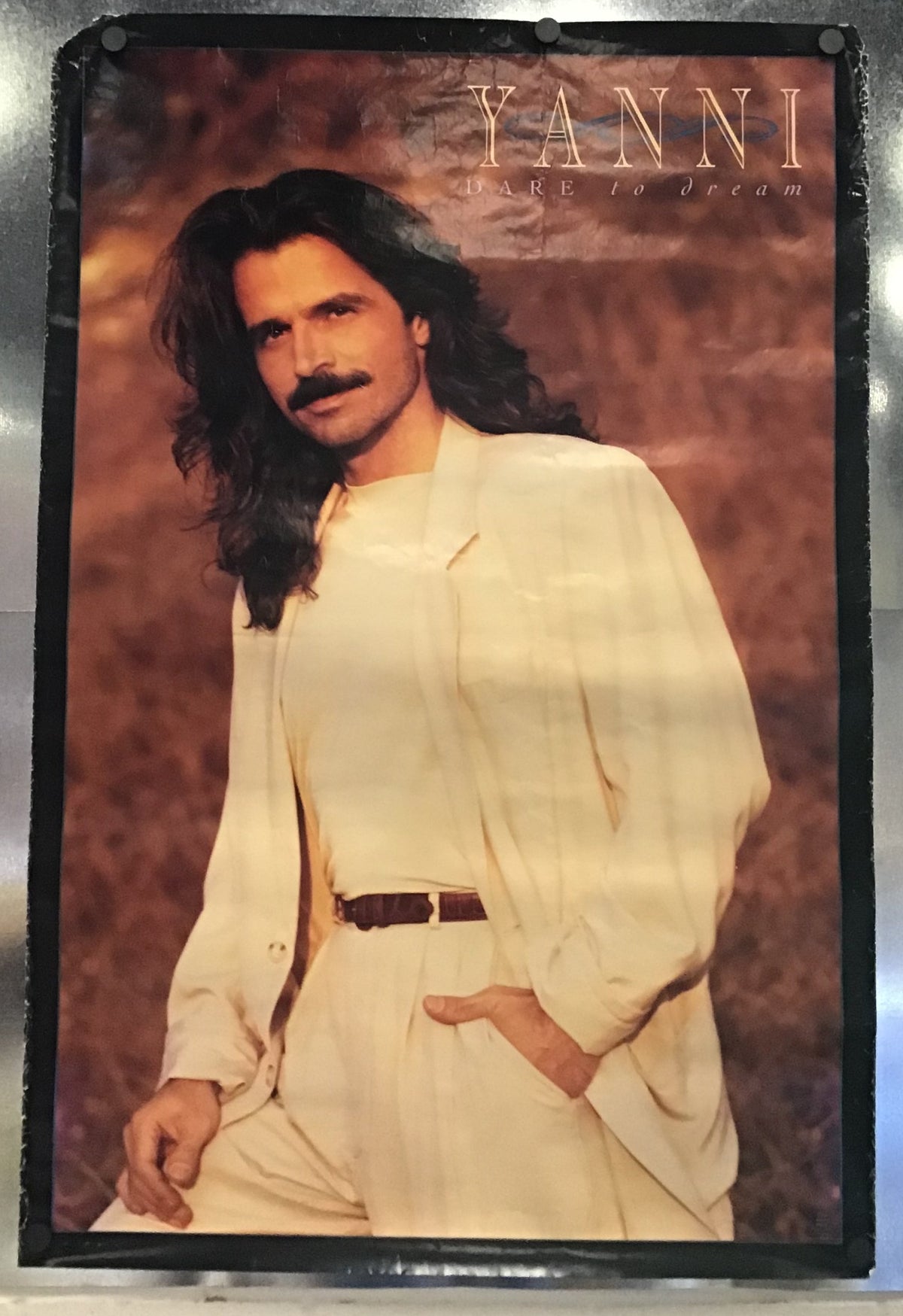 A vintage Yanni 'Dare to Dream' poster featuring the artist with long dark hair and a mustache, wearing a cream-colored outfit against a warm brown background, mounted on a metallic silver wall.